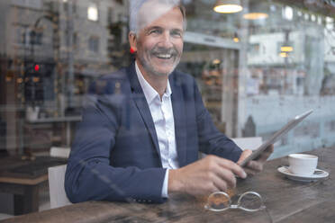 Happy businessman with tablet PC looking through glass window - GUSF07243