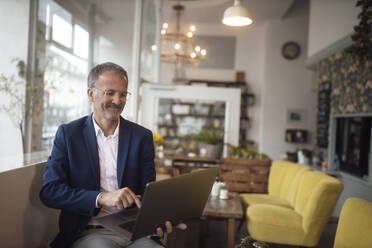 Lächelnder älterer Geschäftsmann mit Laptop in einem Café - GUSF07258