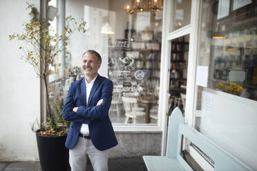 Happy businessman with arms crossed standing in front of glass window - GUSF07262