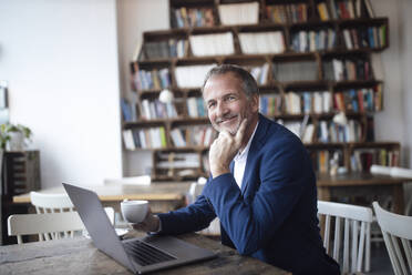 Smiling businessman with coffee cup and laptop sitting at table in cafe - GUSF07263