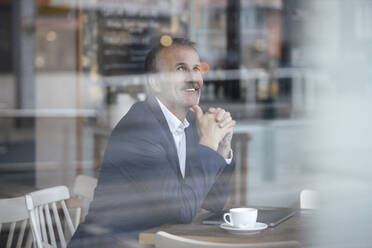 Smiling businessman with coffee cup and laptop seen through glass window at cafe - GUSF07265