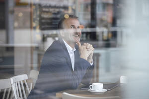 Lächelnder Geschäftsmann mit Kaffeetasse und Laptop, gesehen durch das Glasfenster eines Cafés - GUSF07265