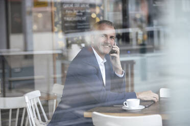 Smiling businessman talking on mobile phone seen through glass window at cafe - GUSF07266