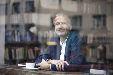Lächelnder Geschäftsmann mit Kaffeetasse durch das Fenster eines Cafés gesehen - GUSF07273