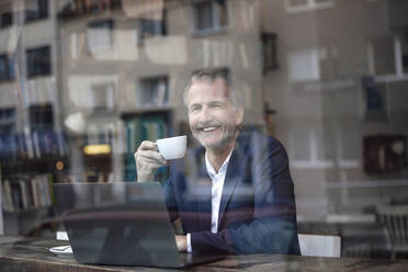 Happy freelancer with coffee cup and laptop seen through glass window at cafe - GUSF07276