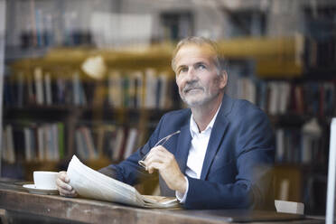 Thoughtful businessman with eyeglasses and newspaper sitting at table in cafe - GUSF07278