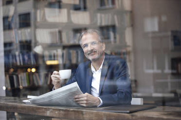 Lächelnder Geschäftsmann mit Kaffeetasse und Zeitung am Tisch im Café sitzend - GUSF07279