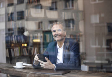 Smiling businessman with smart phone and laptop sitting by glass window at cafe - GUSF07280