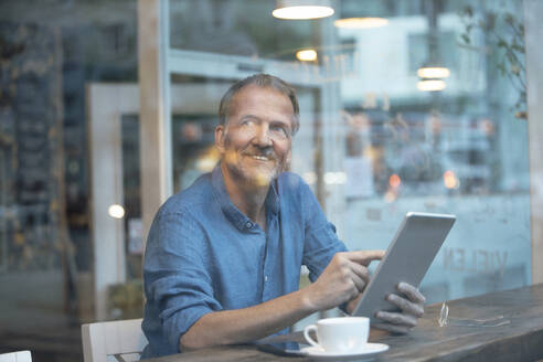 Lächelnder Geschäftsmann mit Tablet-PC, der am Tisch am Fenster in einem Café sitzt - GUSF07286