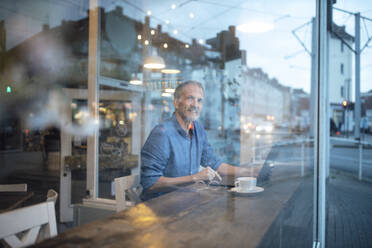 Smiling freelancer with laptop seen through glass window at cafe - GUSF07298