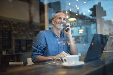Lächelnder Freiberufler mit Laptop, der mit seinem Smartphone spricht, gesehen durch ein Glasfenster in einem Cafe - GUSF07299