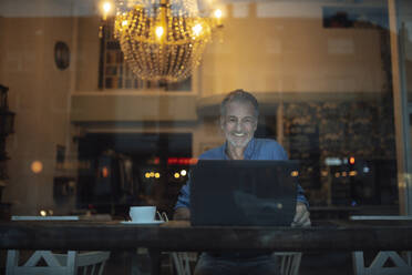 Lächelnder älterer Freiberufler bei der Arbeit am Laptop in einem beleuchteten Café bei Nacht - GUSF07301