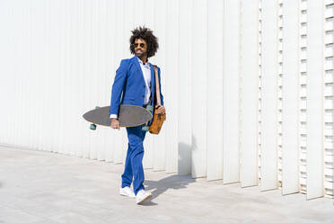 Businessman holding skateboard and shoulder bag walking on footpath - OIPF01410