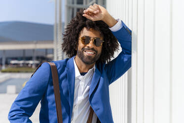 Smiling businessman with curly hair standing by wall on sunny day - OIPF01421