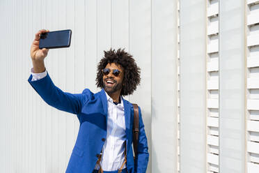 Glücklicher Geschäftsmann, der ein Selfie mit seinem Mobiltelefon macht und vor einer Wand steht - OIPF01425