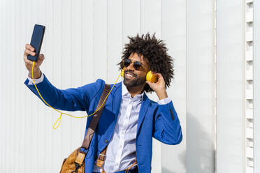 Smiling businessman listening music through headphones taking selfie with mobile phone in front of wall - OIPF01426