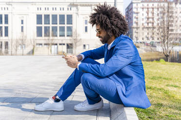 Businessman with curly hair using mobile phone sitting on footpath - OIPF01431