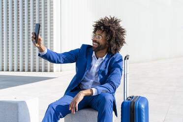 Glücklicher Geschäftsmann mit Koffer, der ein Selfie mit seinem Mobiltelefon vor einer Wand macht - OIPF01434