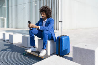 Geschäftsmann mit Koffer und Skateboard, der ein Mobiltelefon benutzt und auf einem Betonblock sitzt - OIPF01436