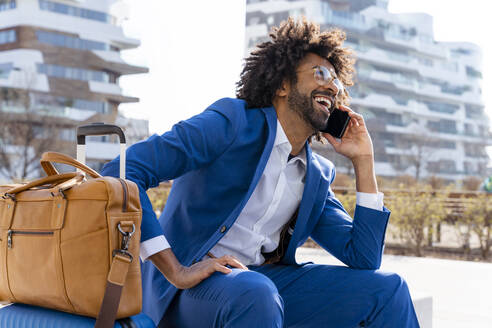 Happy businessman with suitcase and shoulder bag talking on smart phone sitting on concrete block - OIPF01439