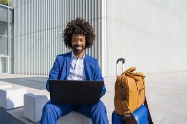 Glücklicher Geschäftsmann, der einen Laptop benutzt und an einem sonnigen Tag neben seinem Gepäck sitzt - OIPF01442
