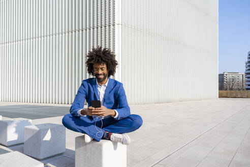 Lächelnder Geschäftsmann, der Musik hört und sein Smartphone benutzt, sitzt auf einem Betonblock - OIPF01454