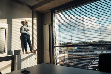 Businesswoman with hand in pocket standing on sideboard in office enjoying the sun - JOSEF08310