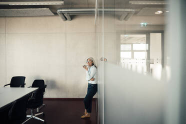 Businesswoman sending voicemail through smart phone leaning on glass wall in office - JOSEF08314