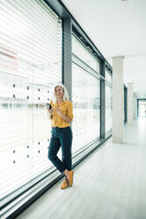 Cheerful businesswoman with smart phone leaning on glass wall in office - JOSEF08317