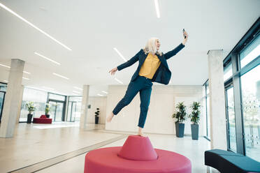 Glückliche Geschäftsfrau nimmt Selfie durch Handy balancieren auf Sitz im Büro - JOSEF08339