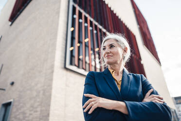 Gray haired businesswoman with arms crossed standing in front of office building - JOSEF08348