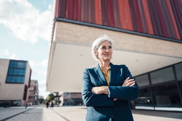 Smiling businesswoman standing with arms crossed outside office building on sunny day - JOSEF08353