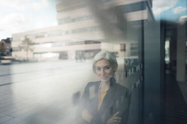 Smiling businesswoman with arms crossed seen through glass standing at office park - JOSEF08357