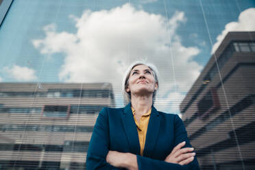 Smiling businesswoman standing with arms crossed in front of glass wall - JOSEF08358