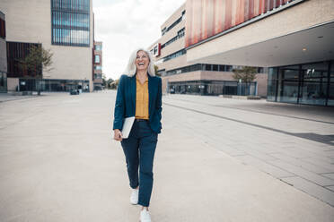 Cheerful businesswoman holding laptop walking with hand in pocket walking at office park - JOSEF08366
