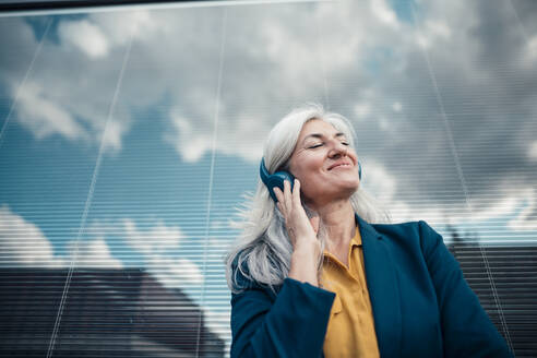 Lächelnde Geschäftsfrau mit geschlossenen Augen, die über drahtlose Kopfhörer vor einer Glaswand Musik hört - JOSEF08370