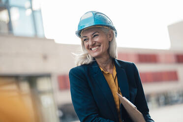 Cheerful female architect with clipboard wearing hardhat at office park - JOSEF08375