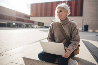 Smiling businesswoman with laptop sitting on seat at office park - JOSEF08385