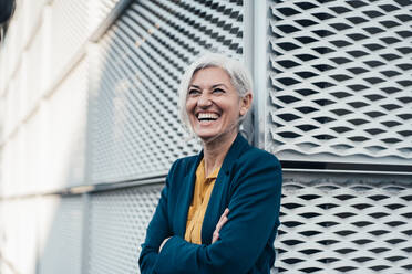 Cheerful businesswoman standing with arms crossed in front of wall - JOSEF08388