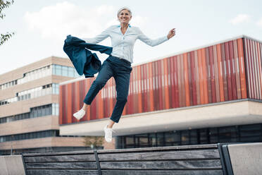 Happy businesswoman with blazer jumping at office park - JOSEF08391