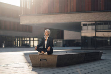 Businesswoman sitting with cross-legged on bench meditating at office park - JOSEF08397