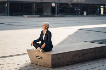 Businesswoman sitting with cross-legged on bench at office park - JOSEF08398