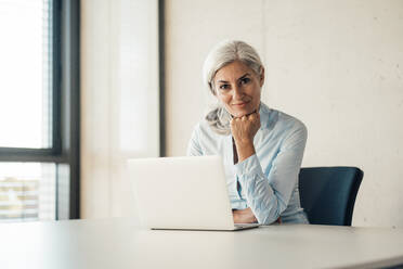 Smiling businesswoman with laptop sitting at desk in office - JOSEF08411