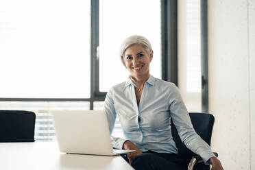 Happy businesswoman with laptop sitting at desk in office - JOSEF08412