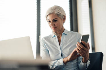 Businesswoman holding smart phone looking at laptop sitting in office - JOSEF08413