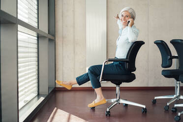 Glückliche Geschäftsfrau, die auf einem Stuhl im Büro sitzt und mit einem Smartphone spricht - JOSEF08417