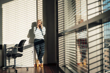 Businesswoman talking on mobile phone standing by glass window in office - JOSEF08418