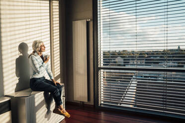 Businesswoman with coffee cup sitting on table looking through glass window - JOSEF08428