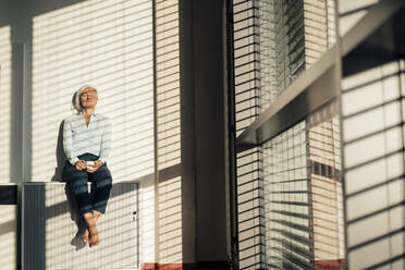 Relaxed businesswoman holding coffee cup sitting with eyes closed on table in office - JOSEF08429