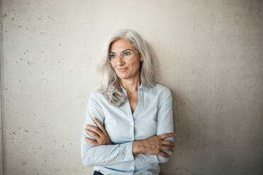 Smiling businesswoman wearing eyeglasses standing with arms crossed in front of wall - JOSEF08438